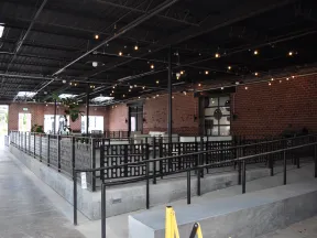 Modern indoor dining area with brick walls, metal railings, string lights, and concrete flooring in an industrial style.