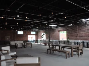 Spacious indoor lounge with wooden furniture, exposed brick walls, and string lights under a high ceiling.