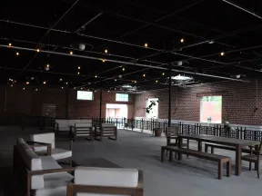 Spacious indoor patio with wooden sofas and tables, exposed brick walls, and string lights overhead.