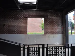 Interior view of an industrial-style building with brick walls, a square window, black railing, and concrete lower wall.