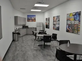 Modern office break room with round tables, black chairs, kitchenette, fridge, and motivational wall posters.
