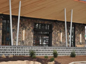 Modern building exterior with stone wall, large windows, vertical lights, and wooden roof overhang.