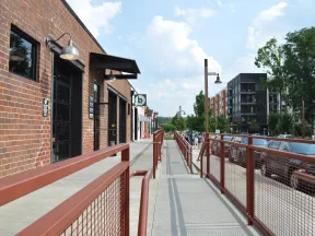 Street view of urban sidewalk with red railings, brick buildings, parked cars, and modern apartments under blue sky.