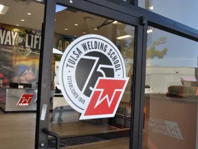 Glass door entrance of Tulsa Welding School with logo and view of interior training area with welding equipment.