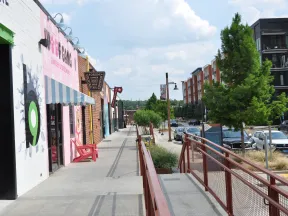 City sidewalk with colorful storefronts, trees, parked cars, and a clear sky on a sunny day