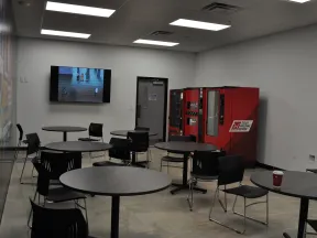 Break room with round tables, black chairs, red vending machines, and a TV mounted on the wall.