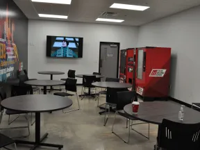 Modern break room with round tables, chairs, vending machines, hand sanitizer, and a TV mounted on the wall.