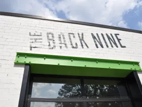 Exterior of a white brick building with The Back Nine sign and green metal awning under a partly cloudy sky
