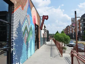 Street view of a colorful mural with geometric trees and rainbow over building, sidewalk, and modern apartments in background