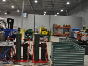 Industrial warehouse with gas cylinders, machinery, storage racks, and plastic containers under bright lighting.