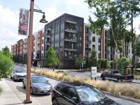 Modern apartment complex with parked cars, street lamps, trees, and landscaped sidewalks on a sunny day.