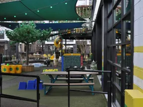 Outdoor play area with picnic tables, green shade sails, playground equipment, and large connect four game board