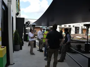 Group of people, including one in a safety vest, talking under a large black canopy outside modern buildings on a sunny day