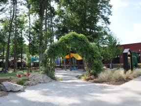Green leafy archway at the entrance of a playground with trees, benches, and a brick building in the background.