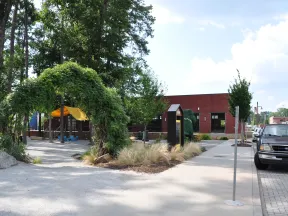 Outdoor park area with green leafy arch, playground with yellow canopy, sidewalk, and parked vehicles on a sunny day
