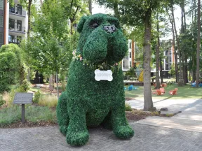 Large topiary sculpture of a dog wearing a floral collar and bone-shaped tag in an urban park setting.
