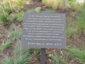 Memorial plaque honoring Scott Selig's vision, empathy, and inspiring life surrounded by plants and grass.