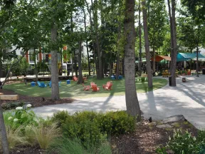 Outdoor urban park with trees, colorful chairs on green lawn, winding pathways, and shaded seating areas under canopies.