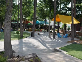Shaded outdoor seating area with colorful chairs and canopies among trees on gravel and grass paths.