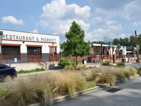 Outdoor view of a restaurant and market with cars, trees, and decorative grasses under a blue sky with clouds