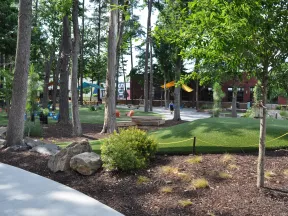 Community park with large trees, green turf, walking paths, playground equipment, and seating areas under sunny sky.