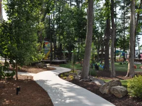 Curved concrete path winding through a wooded outdoor park area with trees, rocks, and picnic tables in sunlight