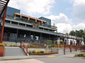 Modern restaurant building with outdoor seating, stairs, ramps, and flower beds under partly cloudy sky.