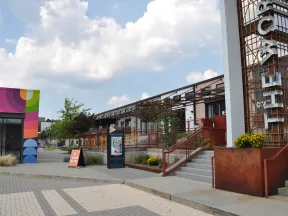 Outdoor urban plaza with colorful mural, modern buildings, stairway, and clear sky with scattered clouds.