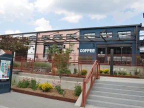 Outdoor view of a coffee shop with seating, plants, stairs, and a colorful directory sign on a sunny day.