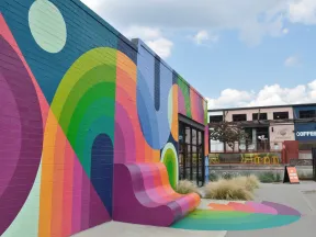 Colorful mural with vibrant rainbow patterns and curved bench on an urban building exterior under blue sky.