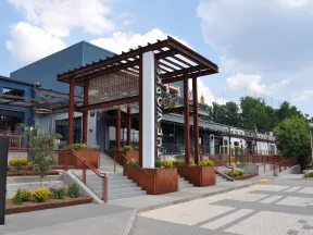 Modern building entrance with rust-colored steel pergola, concrete stairs, and landscaped planters under a partly cloudy sky