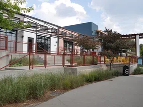 Modern building exterior with red railings, outdoor seating, trees, and a clear sky with clouds.