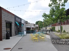 Outdoor seating with colorful chairs and tables along a sidewalk at a cafe under string lights on a sunny day.