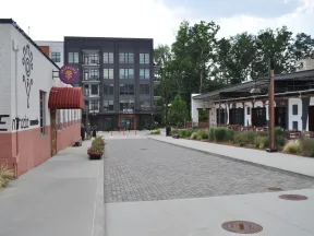 Empty urban street with modern buildings, outdoor seating, greenery, and a cloudy sky background.