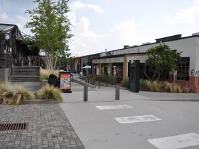 Outdoor shopping and dining area with benches, greenery, and storefronts under a partly cloudy sky in daytime.