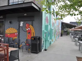 Outdoor seating area with mural of cartoon burger and hot dog high-fiving on brick wall at restaurant.
