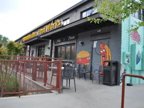 Exterior of a restaurant with murals of a cheeseburger and hot dog waving, outdoor seating, and a red metal fence.