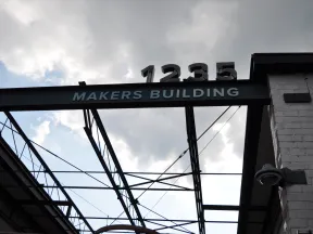 Structural metal beam with Makers Building sign and 1235 number against cloudy sky background.