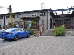 Blue sedan parked in front of a gray industrial Makers Building with stairs and greenery on a cloudy day