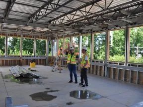 Two construction workers in safety gear discuss plans inside a metal-framed building under construction with large windows.