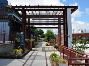 Modern outdoor walkway with metal pergola, planters, and urban buildings under a blue sky with clouds.