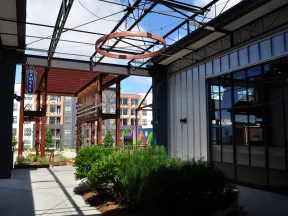 Modern industrial-style outdoor corridor with greenery, metal framework, glass windows, and apartment buildings in background