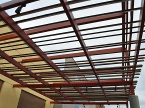 Modern pergola with wooden slats and red beams over a building exterior under cloudy sky.