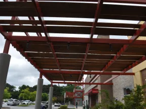 Outdoor walkway with red steel pergola and wooden slats next to a brick building under cloudy sky.