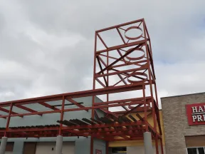 Red metal framework structure with circular cutouts above a building entrance under cloudy sky.