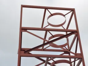 Rust-colored metal framework with circular and diagonal supports against a cloudy sky.