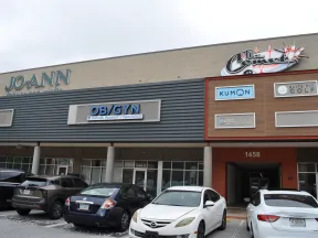 Shopping plaza exterior with storefronts including JOANN, OB/GYN, Kumon, and parking lot with cars in front.