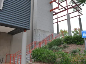 Outdoor staircase with red railings next to modern building and green shrubbery under metal canopy.