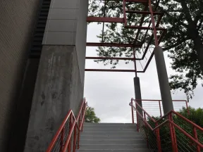 Concrete stairs with red railings lead up to a metal frame structure surrounded by trees against a cloudy sky.