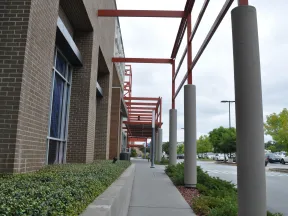 Sidewalk beside a brick building with red steel beams and concrete pillars under an overcast sky near a parking lot.